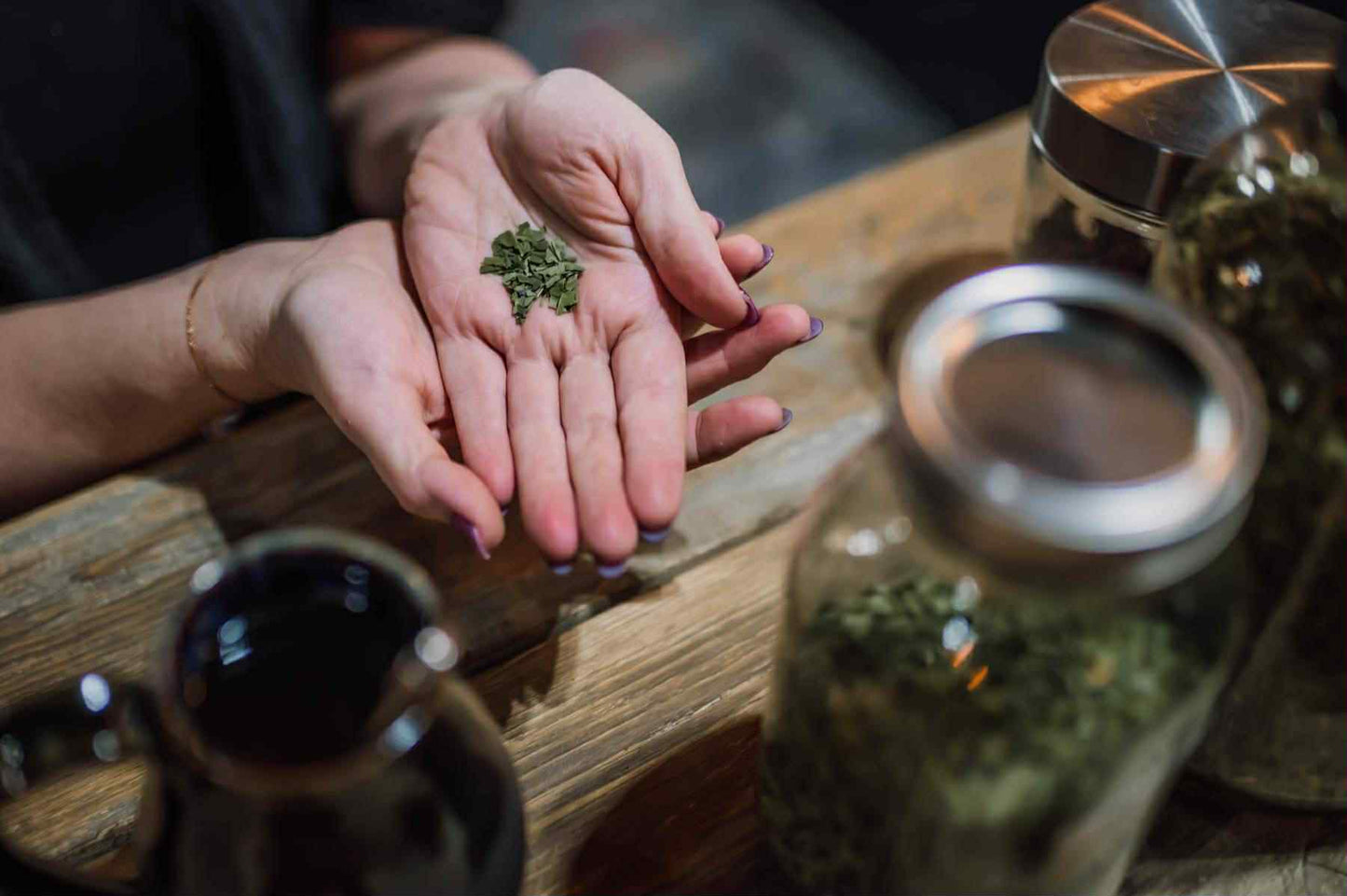 Person holding a small amount of green herbs in their palm on a wooden surface with jars and a teapot. custom tea blending Alberta, herbal tea consultation, Bentley Alberta herbalist, Healing Hills Herbals, personalized tea blend, intuitive herbalism, Central Alberta, Rimbey, Sylvan Lake, Red Deer, Ponoka, Lacombe, Eckville, Edmonton.